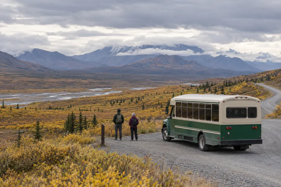 Illustrative shuttle bus and hikers beside a gravel road in autumn tundra