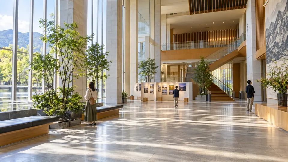 Wide sunlit modern Japanese civic foyer with three visitors looking at architecture and a small exhibition area.