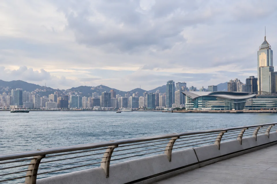 AI-generated illustration of Hong Kong harbour, waterfront railing and a convention venue under an overcast sky.
