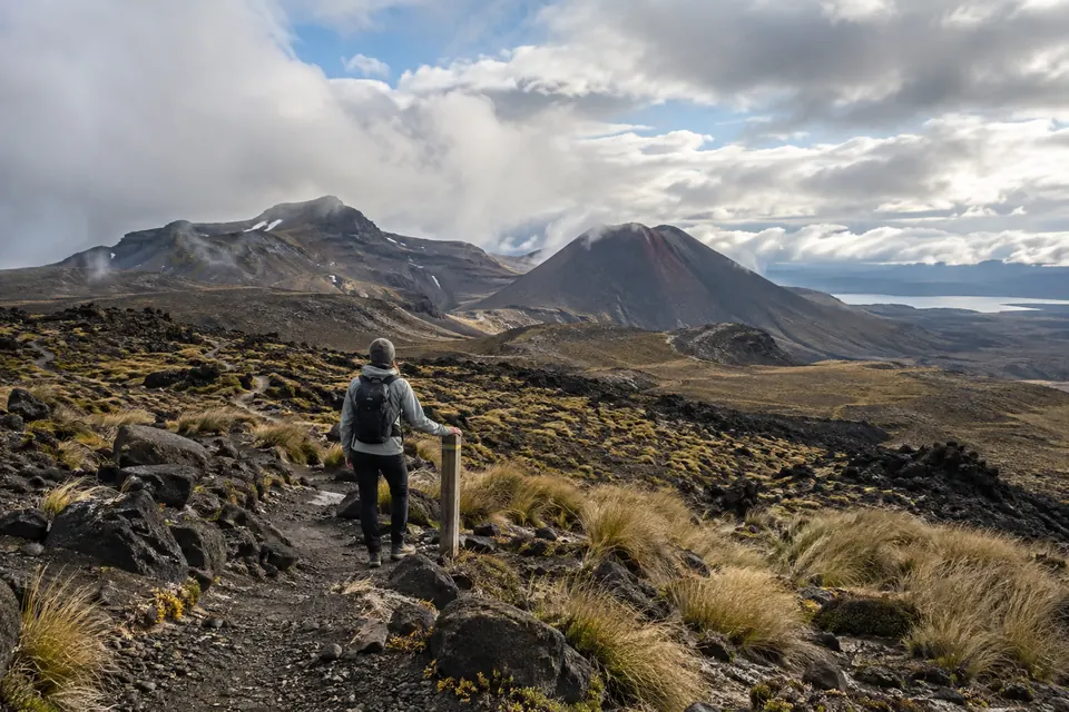 Lone hiker pausing beside an unlabelled route marker on a volcanic alpine trail under changing cloud.