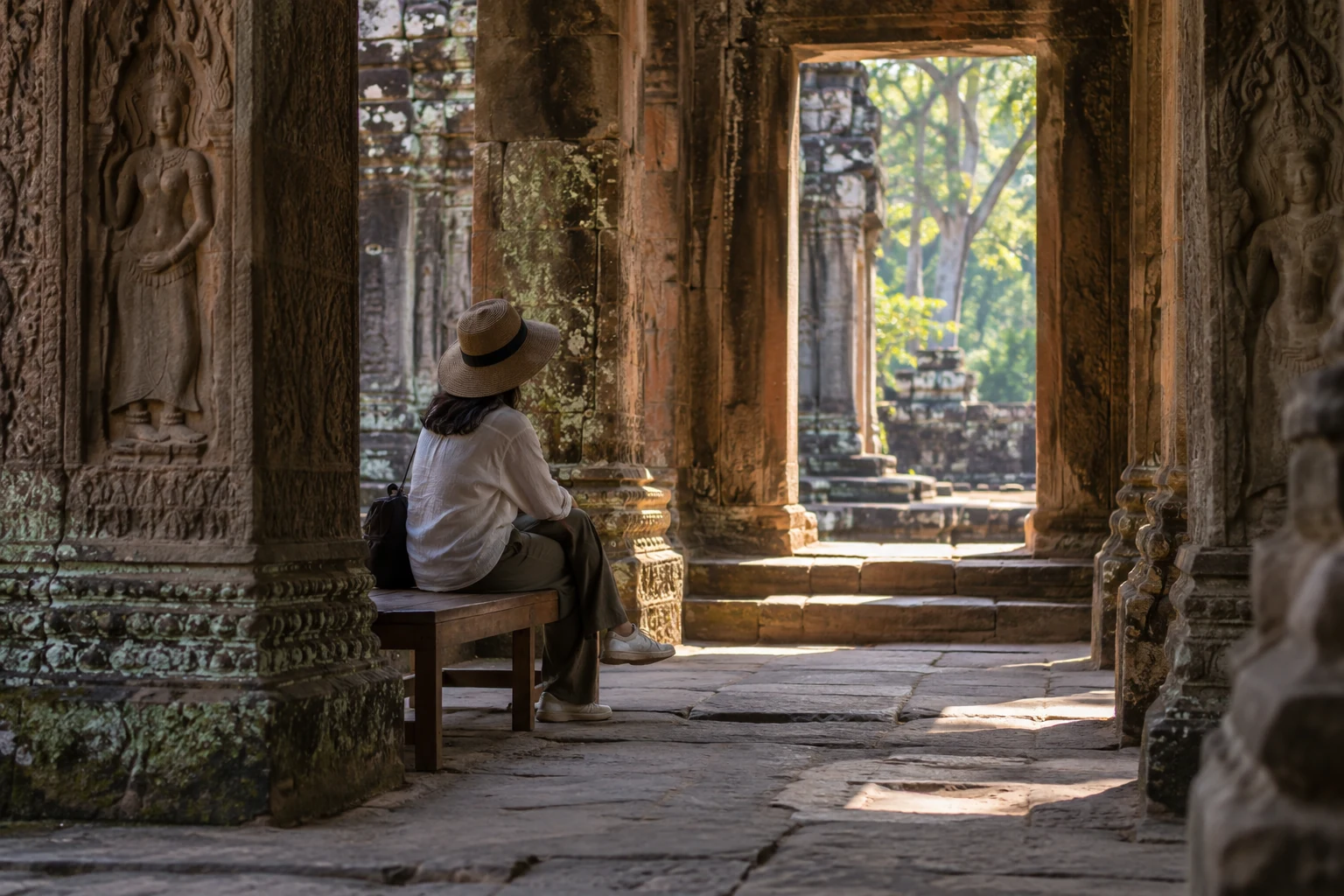 One adult seated on a wooden bench in a shaded carved-stone courtyard.