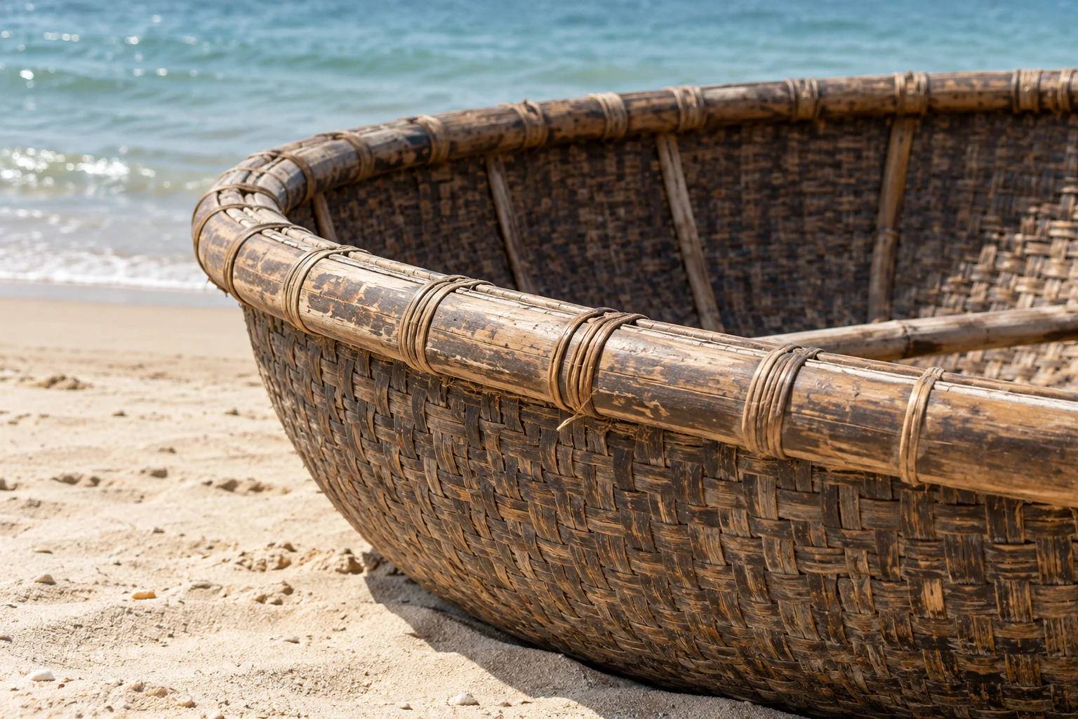 Conceptual close view of a woven round fishing boat on sand beside coastal water.
